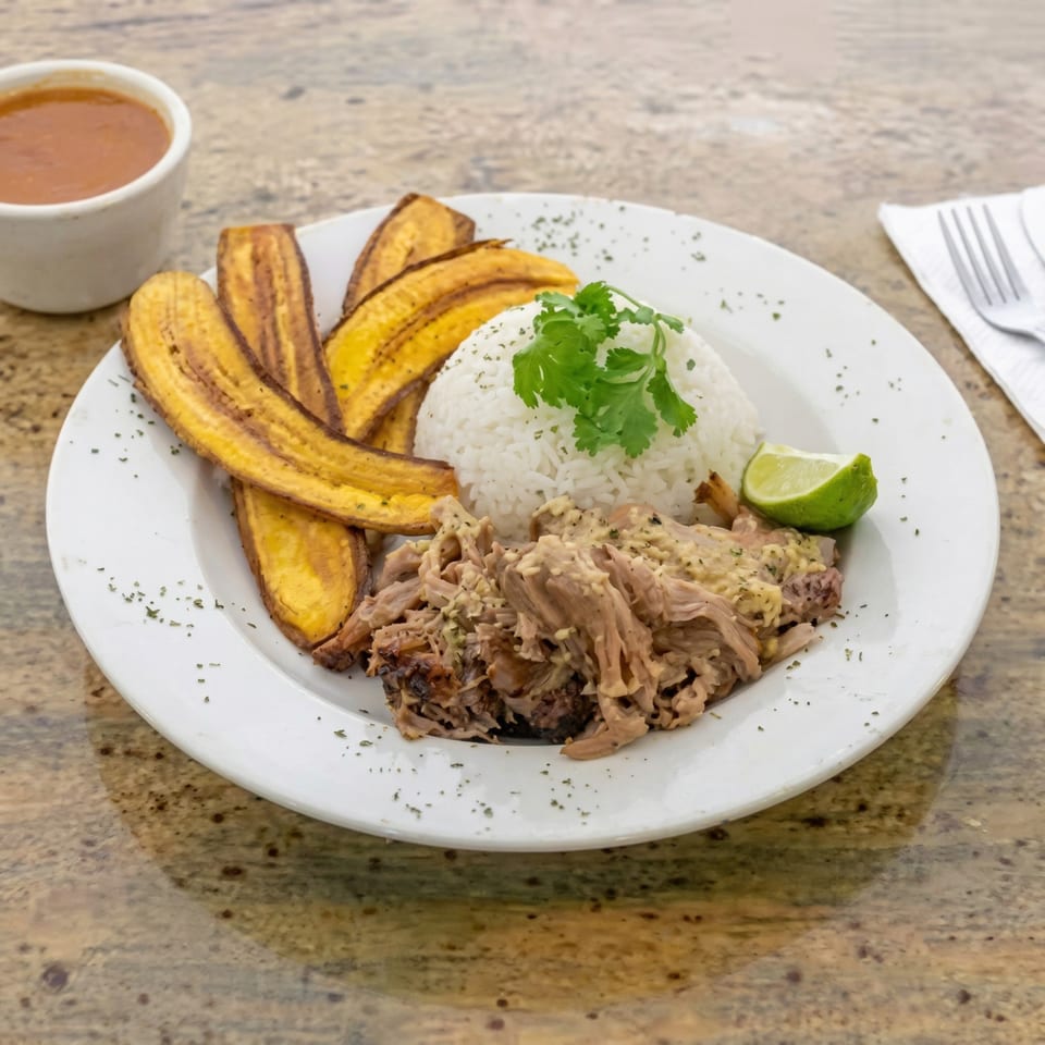 A catering plate with shredded pork, white rice, fried plantains, a lime wedge, and a side of sauce.