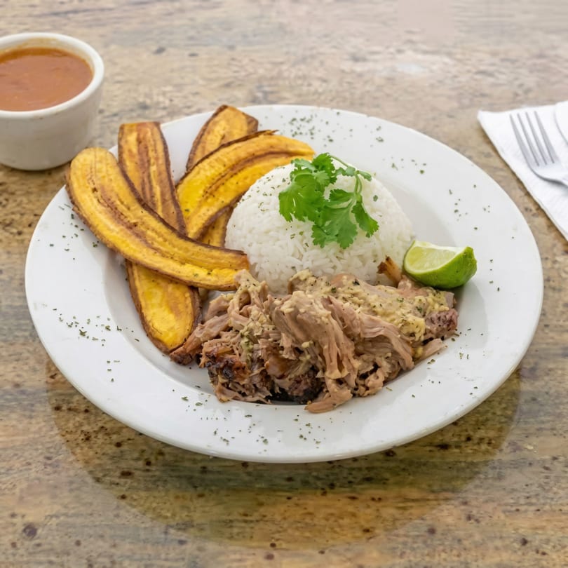 A catering plate with shredded pork, white rice, fried plantains, a lime wedge, and a side of sauce.